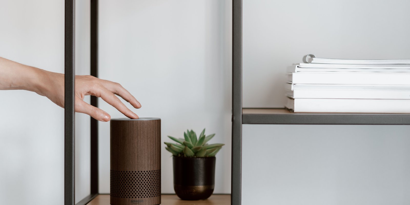 A hand interacts with a smart speaker on a minimalist shelf, featuring a plant and stacked books
