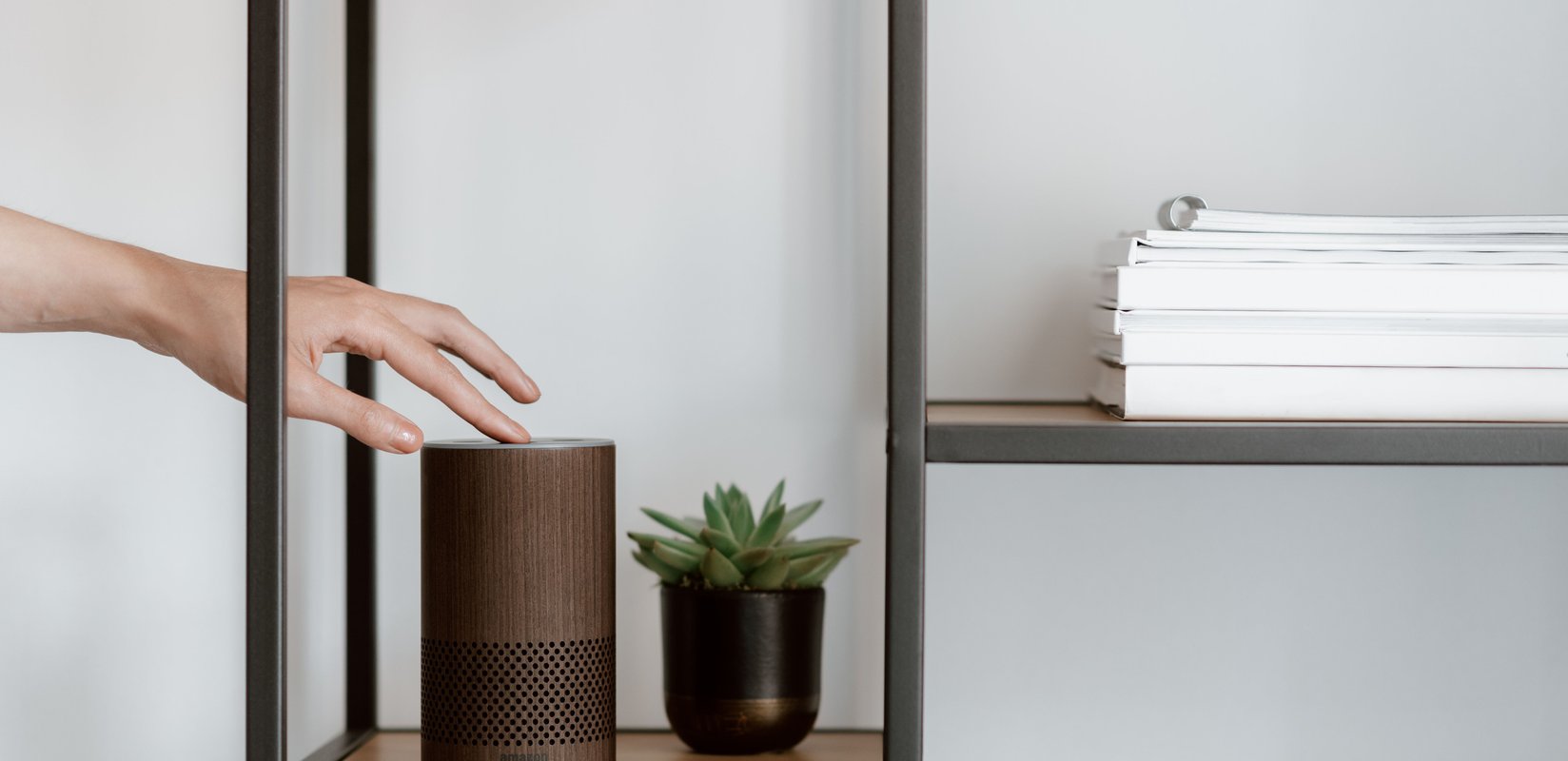 A hand interacts with a smart speaker on a minimalist shelf, featuring a plant and stacked books